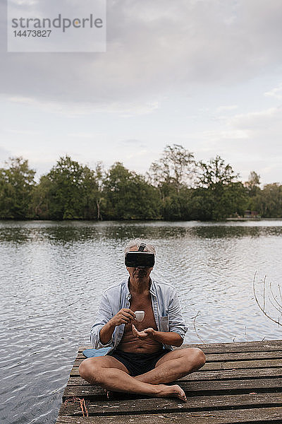 Älterer Mann sitzt auf einem Steg an einem See  trägt eine VR-Brille und trinkt Kaffee