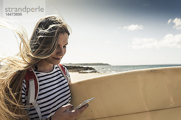Junge Frau am Strand  mit Surfbrett  mit Smartphone