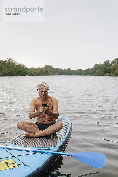 Älterer Mann sitzt mit seinem Handy im SUP-Board auf einem See