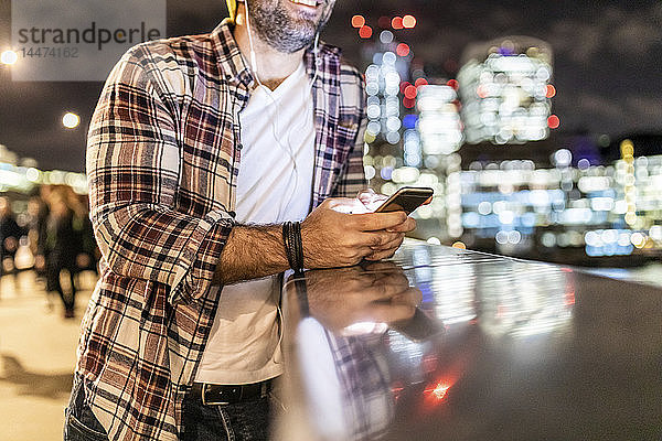 Grossbritannien  London  Nahaufnahme eines an einem Geländer lehnenden Mannes mit Mobiltelefon mit Stadtlichtern im Hintergrund