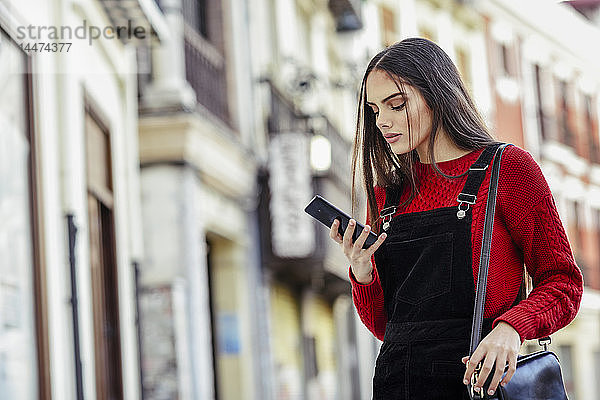 Modische junge Frau mit Blick auf Handy in der Stadt