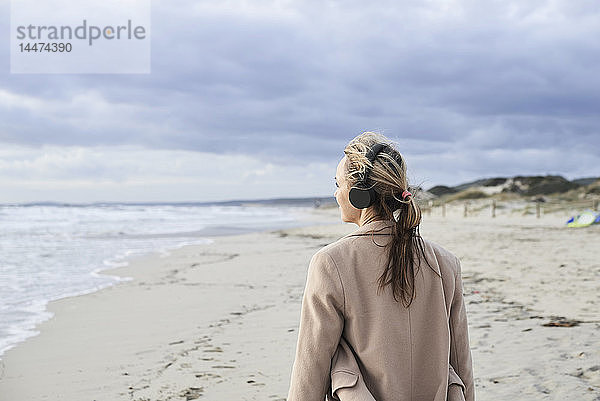Spanien  Menorca  Rückenansicht einer älteren Frau  die im Winter am Strand mit drahtlosen Kopfhörern Musik hört
