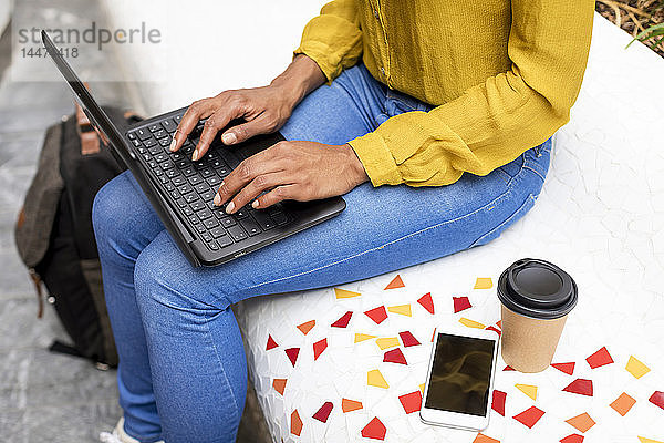 Frau sitzt auf Bank mit Smartphone und Kaffee zum Mitnehmen mit Laptop  Teilansicht