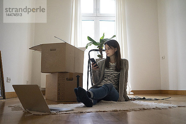 Junge Frau sitzt auf dem Boden ihres neuen Hauses und benutzt ein Smartphone