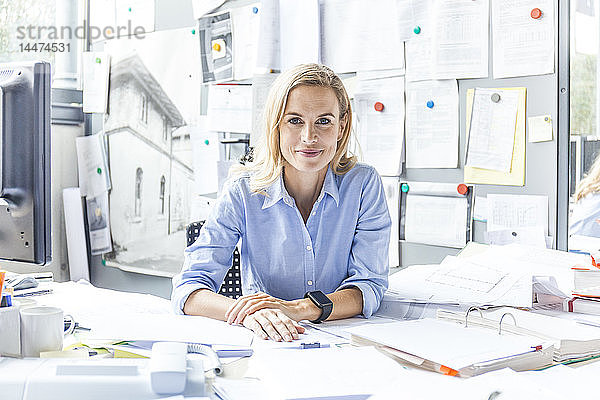 Porträt einer selbstbewussten Frau  die am Schreibtisch im Büro sitzt und von Papierkram umgeben ist