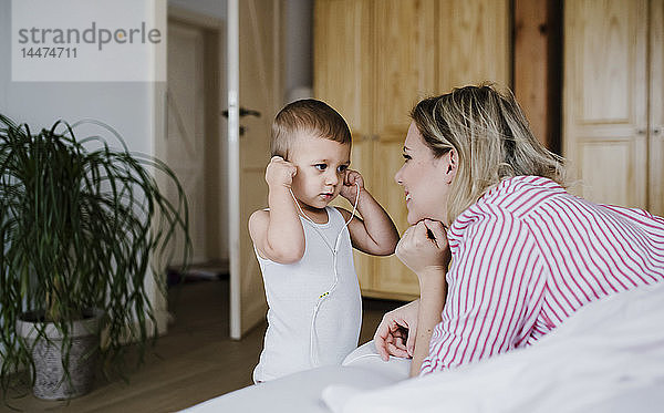 Lächelnde Mutter beobachtet Kleinkind-Sohn mit Kopfhörern im Schlafzimmer zu Hause