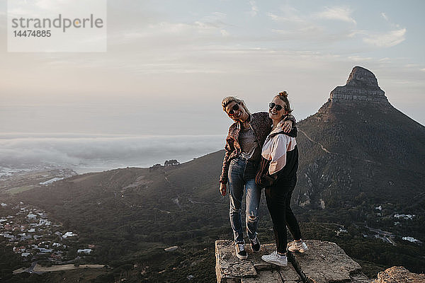 Südafrika  Kapstadt  Kloof Nek  Porträt von zwei glücklichen Frauen  die bei Sonnenuntergang auf einem Felsen stehen