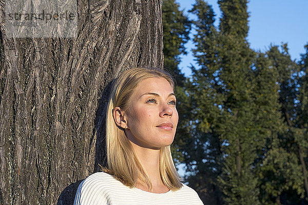 Porträt einer tagträumenden blonden Frau  die sich an einen Baumstamm lehnt und in die Ferne schaut