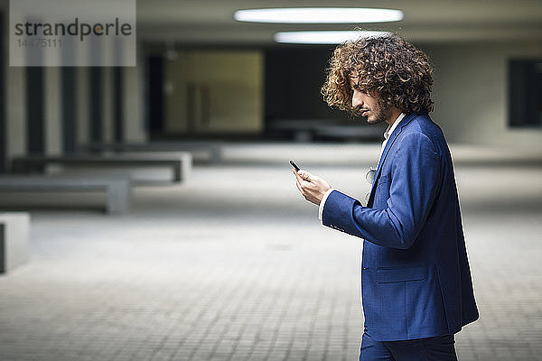 Junger  modischer Geschäftsmann mit lockigem Haar schaut auf Smartphone