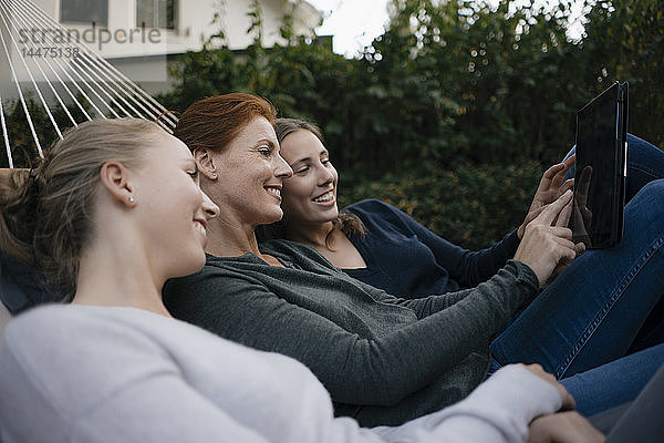 Glückliche Mutter mit zwei Teenager-Mädchen  die im Herbst in der Hängematte im Garten liegen und Tabletten benutzen