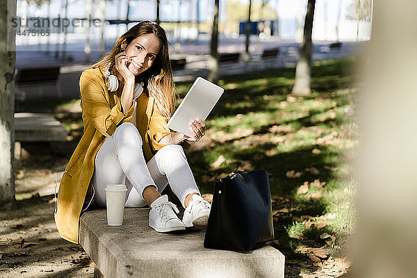 Lächelnde Frau sitzt auf einer Parkbank und benutzt ein Tablett