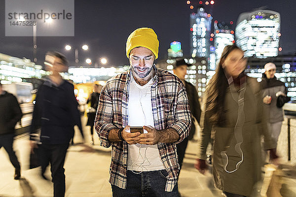 Großbritannien  London  lächelnder Mann  der nachts auf sein Telefon schaut  während in der Nähe verschwommene Menschen vorbeigehen