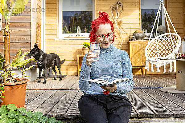 Porträt einer lächelnden älteren Frau mit rot gefärbten Haaren  die auf der Terrasse vor ihrem Haus sitzt und ein Buch liest