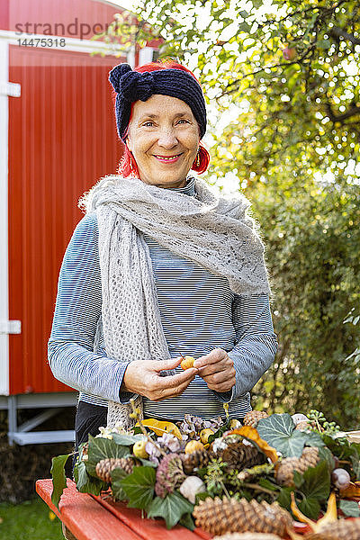 Porträt einer lächelnden älteren Frau mit rot gefärbten Haaren beim Basteln an der Herbstdekoration im Garten