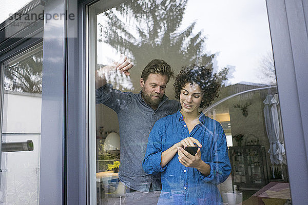 Ehepaar zu Hause hinter dem Fenster mit Mobiltelefon