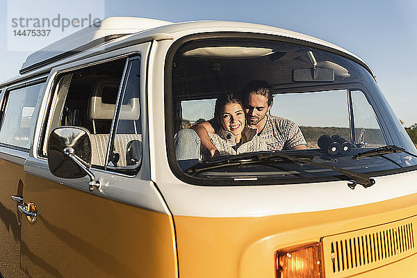 liebevolles Paar  das in seinem Wohnwagen sitzt  die Arme um sich legt und den Sonnenuntergang betrachtet