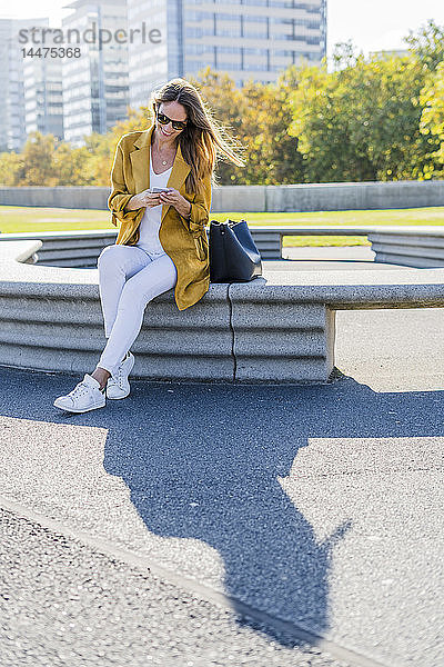 Lächelnde Frau mit Tasche sitzt auf einer Bank in der Stadt und benutzt ihr Handy