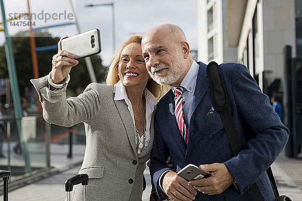 Leitender Geschäftsmann und Geschäftsfrau mit Gepäck  die sich in der Stadt ein Selfie nehmen