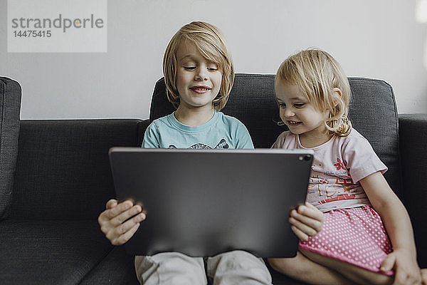 Portrait von Bruder und kleiner Schwester  die zu Hause auf der Couch sitzen und ein digitales Tablet benutzen