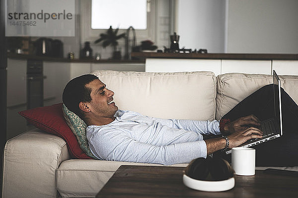 Lächelnder junger Mann  der zu Hause auf dem Sofa liegt und seinen Laptop benutzt