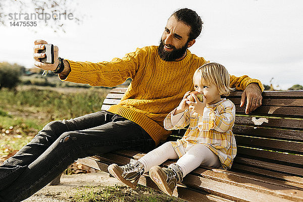 Vater und Tochter sitzen im Herbst auf einer Parkbank  der Vater nimmt ein Selfie  die Tochter isst einen Apfel