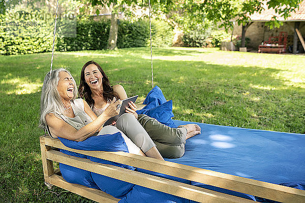 Zwei lachende Frauen entspannen sich mit Tabletten auf einem hängenden Bett im Garten