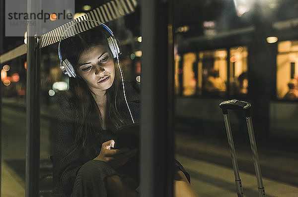 Porträt einer jungen Frau mit Kopfhörern  die nachts auf dem Bahnhof wartet und ein Tablett benutzt