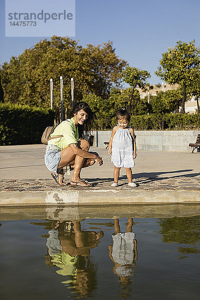 Glückliche Mutter mit Tochter  die ihr Spiegelbild in einem Pool in einem Park betrachten