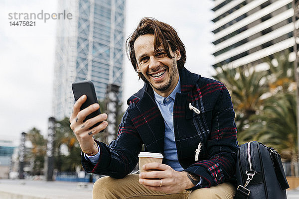 Spanien  Barcelona  glücklicher Mann sitzt in der Stadt mit Kaffee zum Mitnehmen und Handy