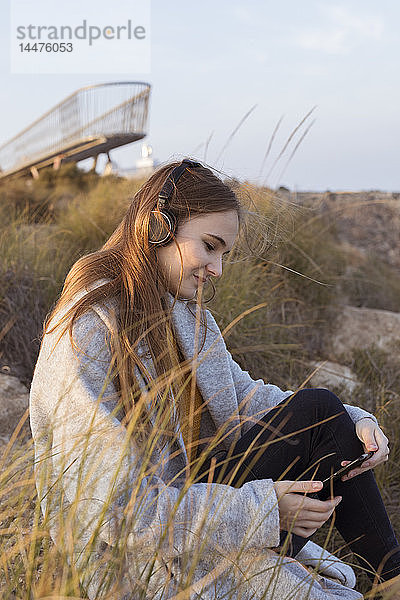 Spanien  Alicante  Santa Pola  Kap Santa Pola  junge Frau sitzt bei Sonnenuntergang auf einem Felsen und hört Musik auf ihrem Handy