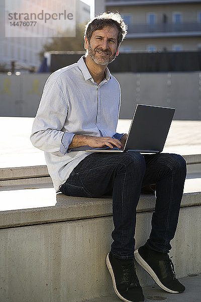Porträt eines lächelnden reifen Mannes  der in der Stadt mit einem Laptop sitzt