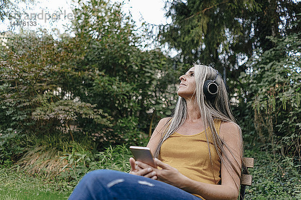 Frau mit Handy sitzt im Garten und hört Musik mit Kopfhörern