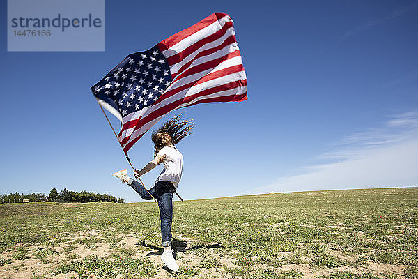 Glückliches Mädchen hält amerikanische Flagge und springt auf Feld in entlegener Landschaft