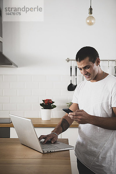 Lächelnder junger Mann benutzt Laptop und Handy in der Küche zu Hause