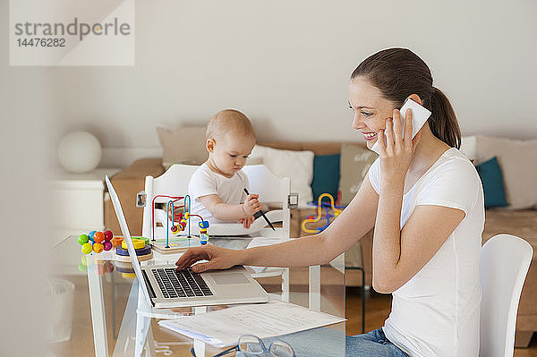 Lächelnde Mutter benutzt Laptop und Handy  während die kleine Tochter zu Hause am Tisch spielt