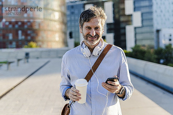 Porträt eines lächelnden reifen Mannes mit Kaffee zum Mitnehmen  Kopfhörer und Mobiltelefon in der Stadt