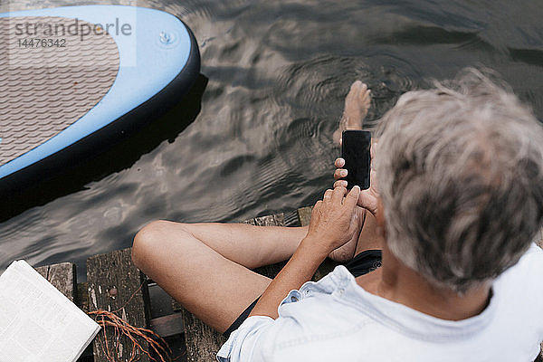 Älterer Mann sitzt auf einem Steg an einem See und benutzt sein Handy