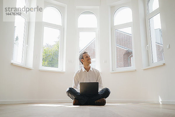 Geschäftsmann sitzt auf dem Boden seines frisch renovierten Hauses  benutzt Laptop und lächelt