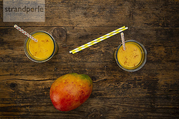 Mango und zwei Gläser Mango Lassi mit Kurkuma und Strohhalmen auf Holz