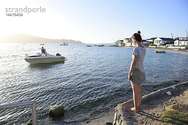 Junge Frau steht an der Küste und sieht einen Mann auf einem Boot im Meer an
