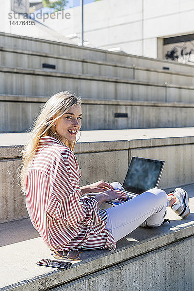 Porträt einer lächelnden jungen Frau mit Laptop  die auf einer Treppe im Freien sitzt und etwas beobachtet