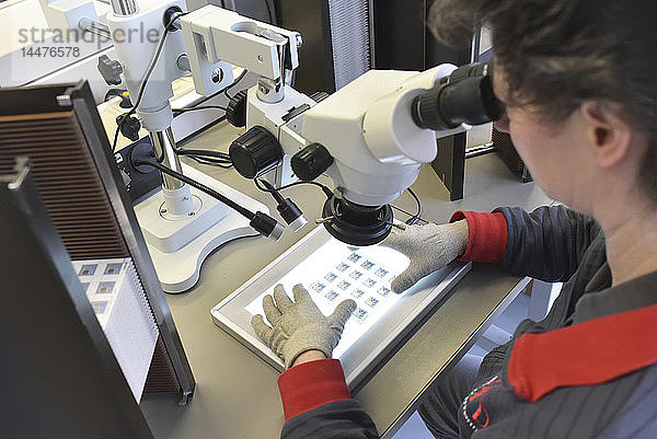 Frau  die ein Mikroskop für die Qualitätskontrolle bei der Herstellung von keramischen Leiterplatten für die Elektronikindustrie benutzt