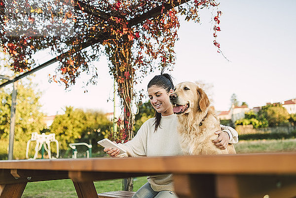 Lächelnde junge Frau mit Handy und ihrem Golden-Retriever-Hund  der sich in einem Park ausruht