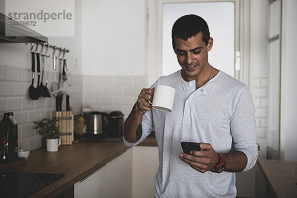 Lächelnder junger Mann mit einer Tasse Kaffee  der sein Handy in der Küche zu Hause benutzt