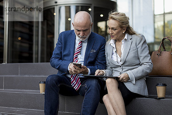 Leitender Geschäftsmann und Geschäftsfrau sitzen auf einer Treppe und benutzen Handy und Tablet