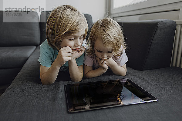 Bruder und kleine Schwester liegen zu Hause auf der Couch und benutzen ein digitales Tablett