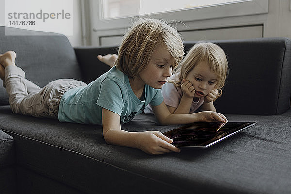 Bruder und kleine Schwester liegen zu Hause auf der Couch und benutzen ein digitales Tablett