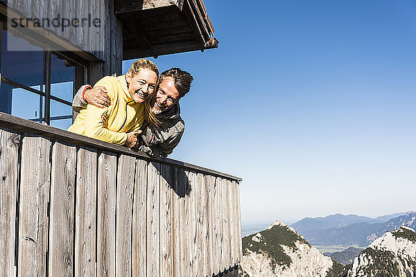 Auf dem Balkon einer Berghütte lehnendes Ehepaar  die Arme um den Kopf gelegt