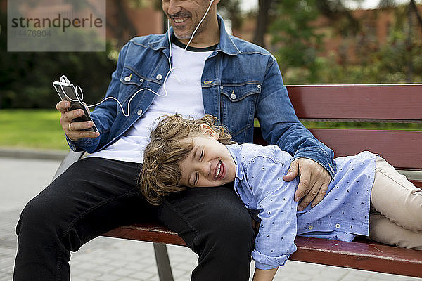 Glücklicher Vater mit Sohn auf einer Bank mit Handy und Ohrstöpseln