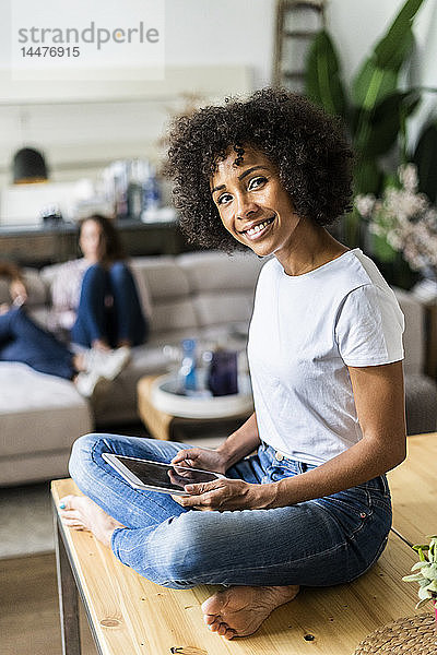 Porträt einer lächelnden Frau mit Tablette auf dem Tisch zu Hause mit Freunden im Hintergrund